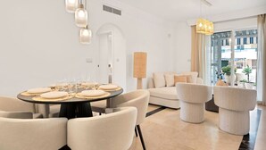 Un espace repas moderne avec une table ronde dressée pour six personnes, entourée de chaises de couleur crème. On aperçoit au fond un canapé avec un coussin assorti, ainsi qu’une grande fenêtre offrant une vue sur la ville.