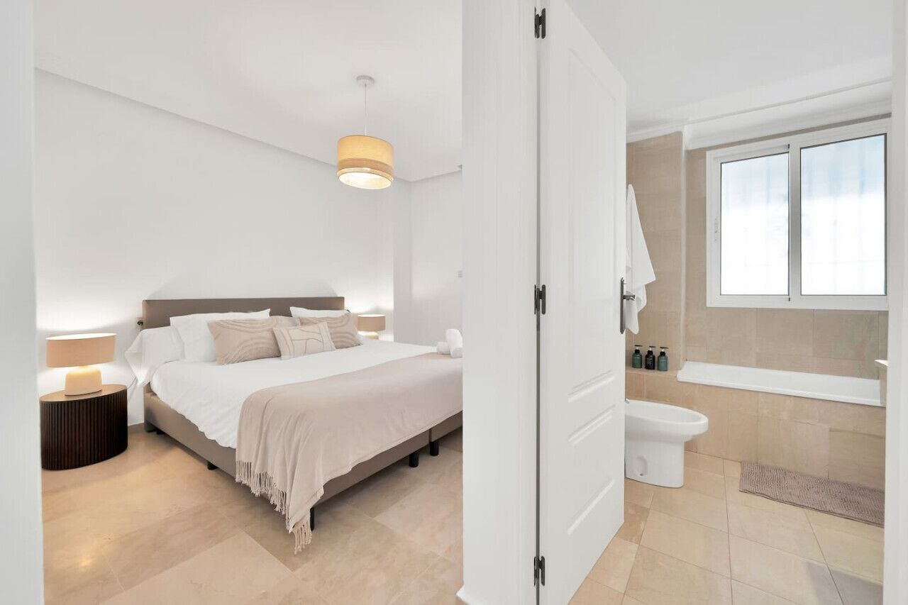 A modern bedroom with a bed, bedside lamps, a nightstand, and a bathroom with a toilet and bathtub.