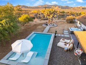 Piscina con sillas de descanso y una sombrilla, rodeada por una zona de patio con vistas a colinas desérticas.