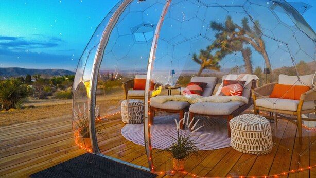 Estructura de cúpula transparente con asientos en su interior, con vistas a un paisaje con árboles y colinas.