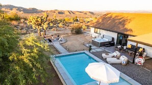 Piscina rectangular con una sombrilla blanca, sillas de descanso y un jacuzzi en un entorno desértico.