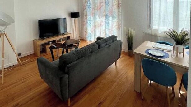 A modern living room with a grey sofa, wooden flooring, a TV on a stand, a dining table with chairs, and a window with curtains.