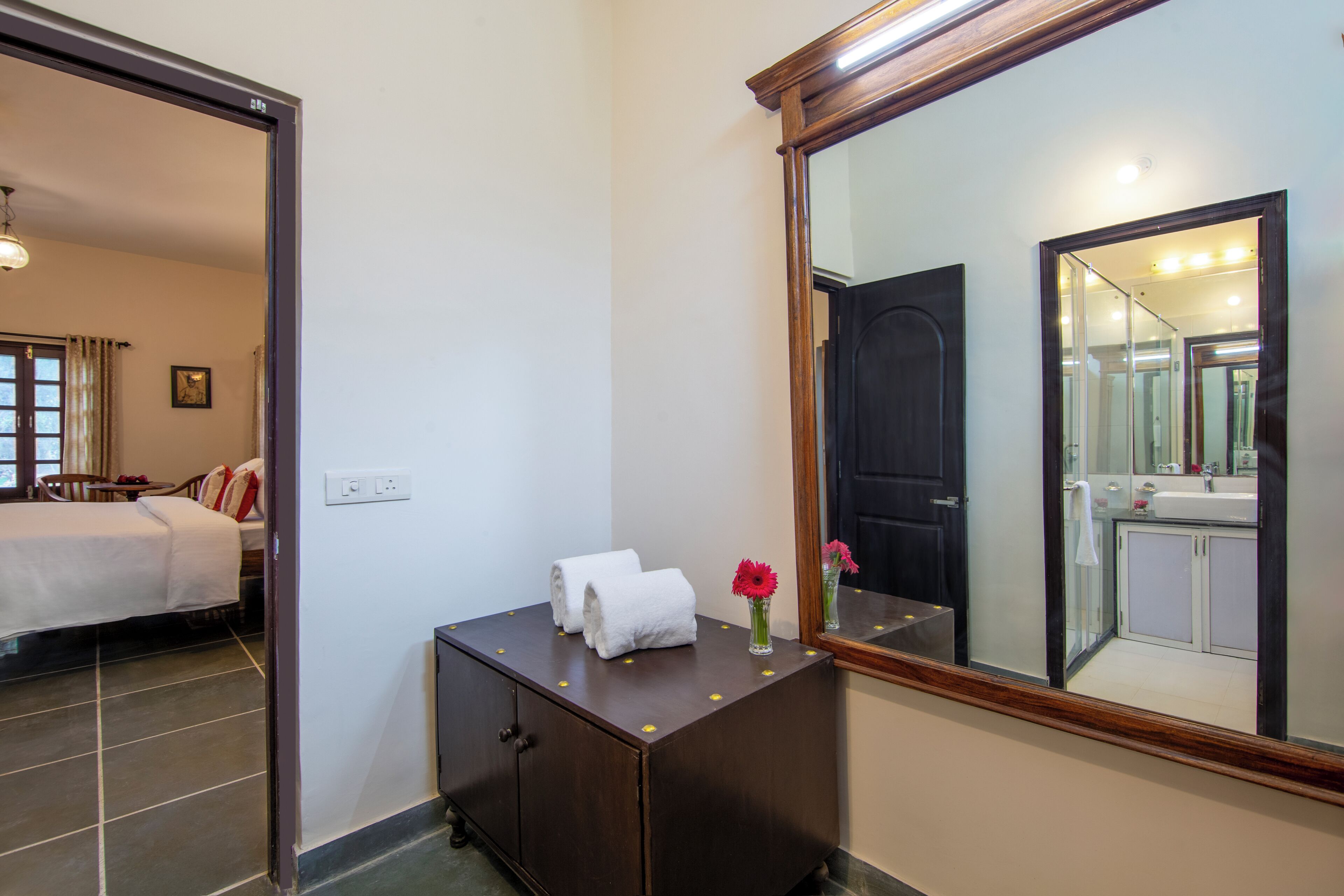 A hotel room with a bed, a wooden dresser with towels and a vase of flowers, a large mirror, and a bathroom visible through an open door.