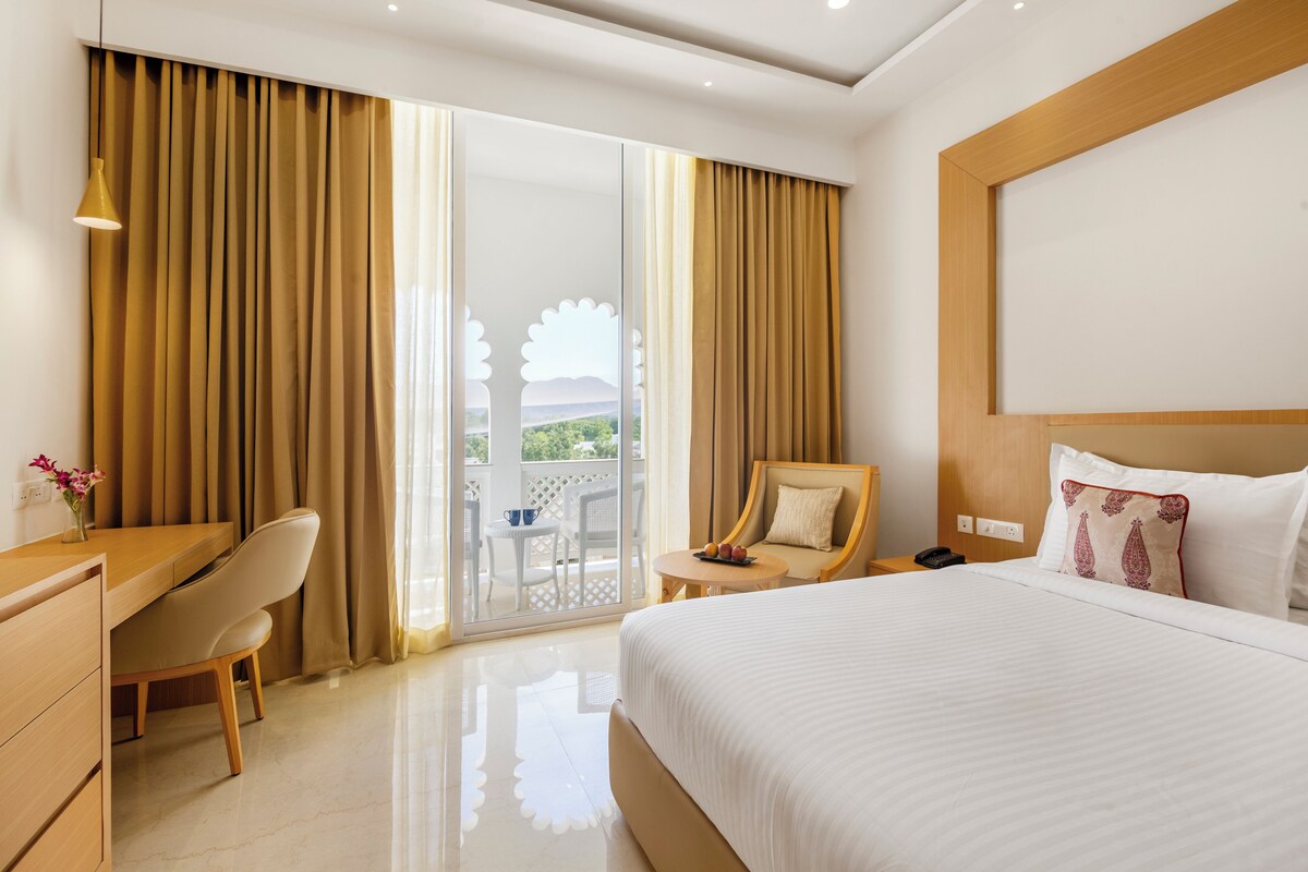 A hotel room with a large bed, a desk, a chair, and a view through a sliding door.
