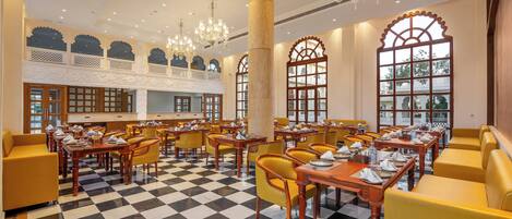 A spacious dining area with checkered flooring, yellow upholstered seating, and chandeliers.