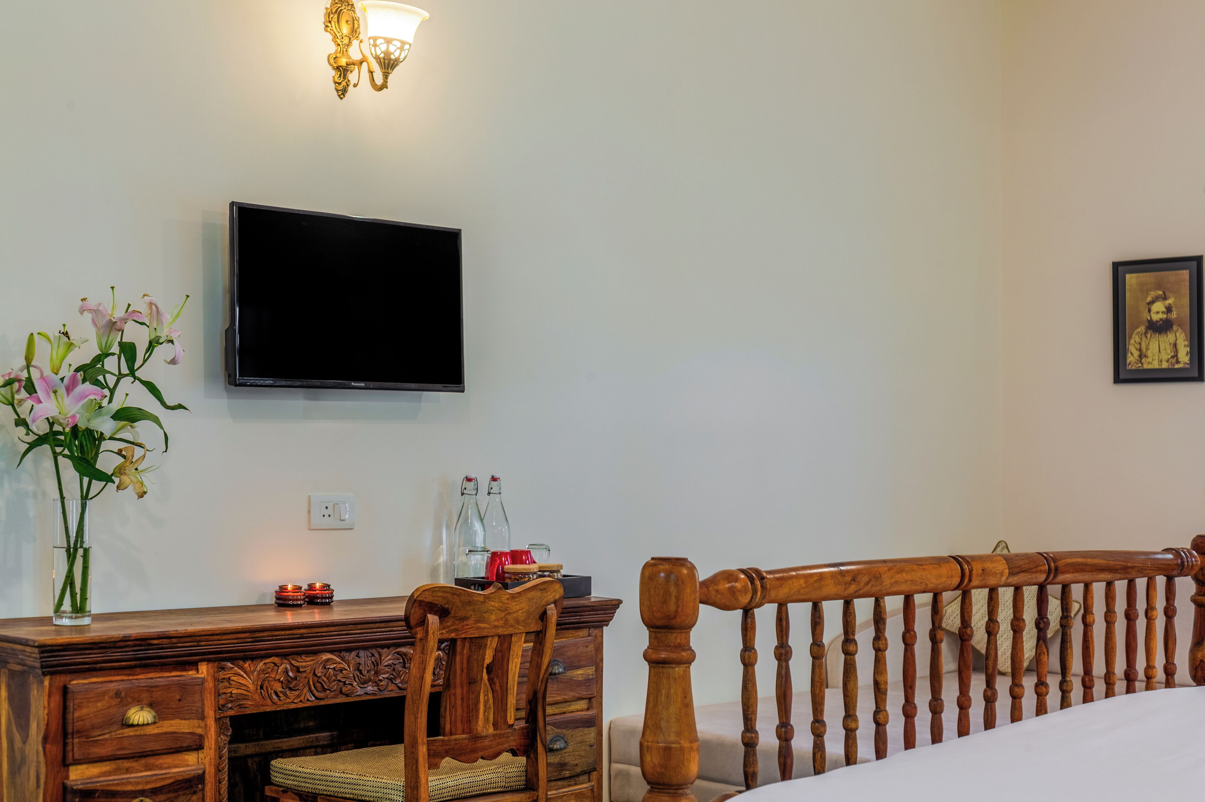 A hotel room with a wooden desk, a television mounted on the wall, a vase with flowers, and a bed with a wooden headboard.
