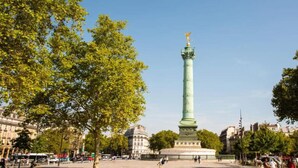 Un alto monumento verde con una statua in cima, circondato da alberi e edifici.