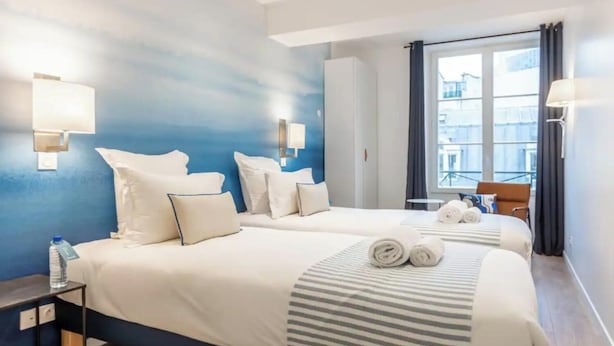 Una camera d'albergo con due letti, vista sugli edifici dalla finestra e una sedia vicino al vetro.