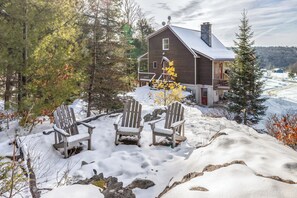 Uma cabana com fachada de madeira, um deck coberto de neve e duas cadeiras Adirondack.