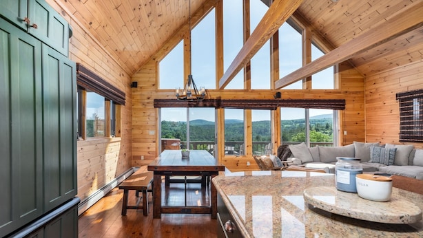 Une cabane en bois spacieuse, dotée d’une grande table à manger, d’un coin cuisine et d’un espace salon offrant une vue sur les montagnes.