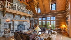 A spacious living room with a stone fireplace, wooden beams, and large windows. - Mountainside 5-BR Ski-In/Out Okemo Villa (Ludlow)