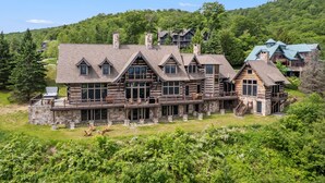 A large log cabin with multiple levels, stone foundations, and a balcony overlooking a lush green landscape. - Mountainside 5-BR Ski-In/Out Okemo Villa (Ludlow)