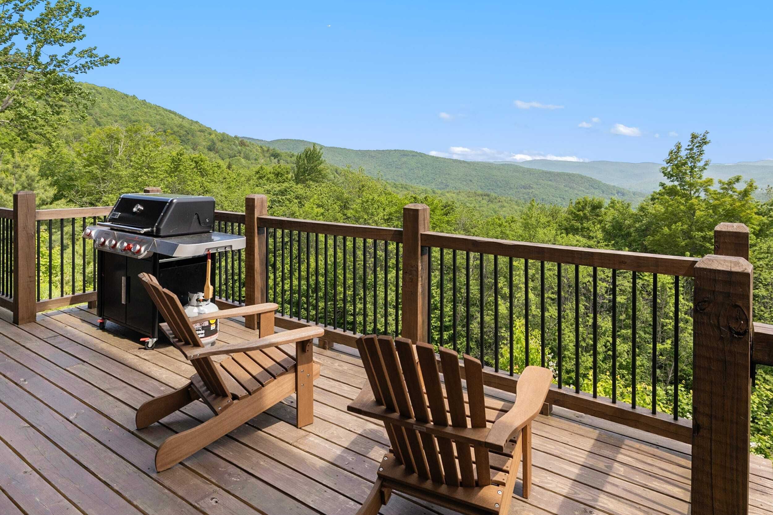 A wooden deck with a grill and two Adirondack chairs overlooking a forested mountain range.