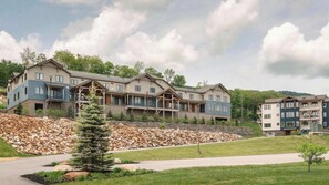 A multi-story residential building with a stone retaining wall, a paved driveway, and a small tree in the foreground. - Ski-In/Out Condo on Okemo Mountain (Windsor County)