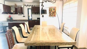 A dining area with a large marble table surrounded by chairs, a kitchen in the background, and a poster on the wall. - Evergreen Resort & Farm Aravali (Gurugram)
