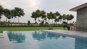 A swimming pool with a tiled deck, surrounded by a grassy area and trees. - Evergreen Resort & Farm Aravali (Gurugram)