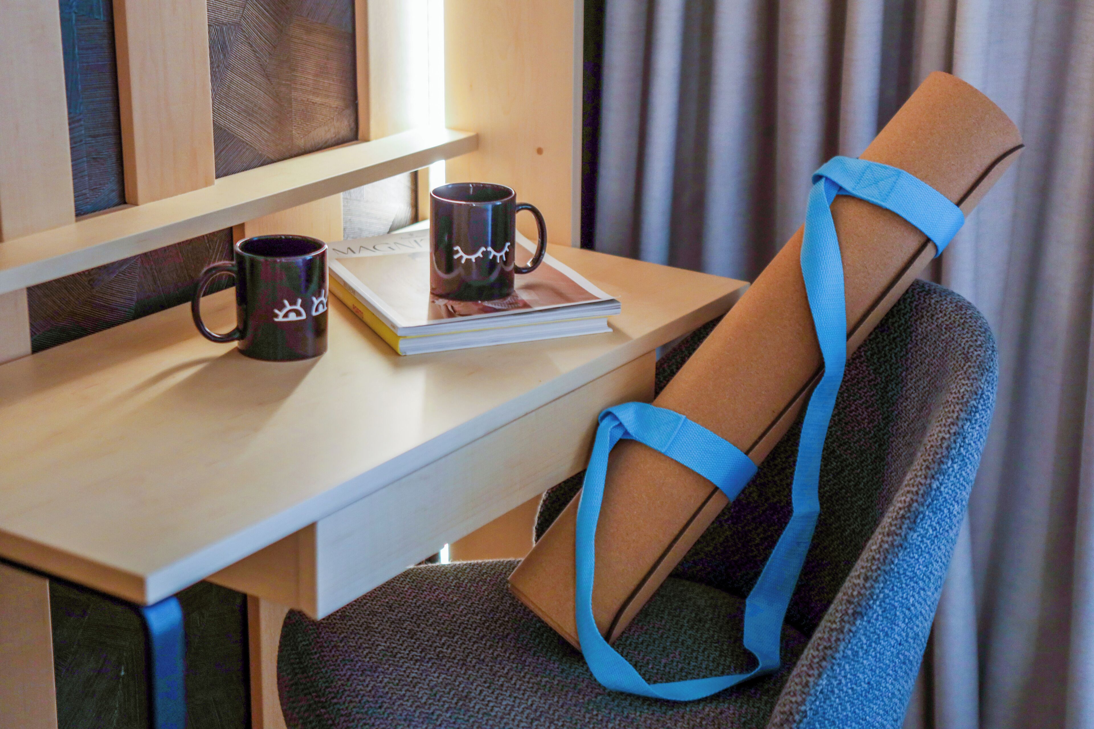 A wooden desk with two mugs and a book, a blue seatbelt, and a window with curtains.