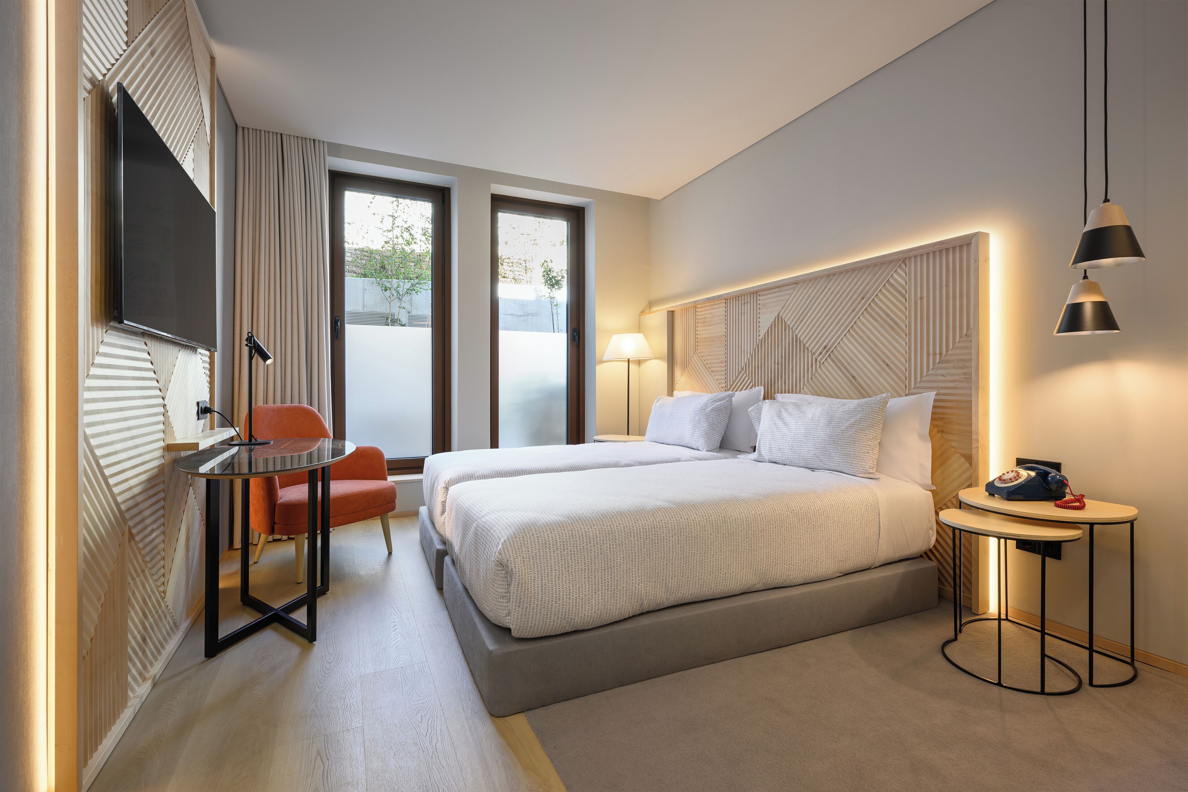 Quarto de hotel moderno com uma cama grande, uma escrivaninha com cadeira, uma TV e uma mesinha de cabeceira com abajur.