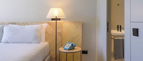 Une chambre d’hôtel avec un lit, une lampe de chevet, une petite table avec un téléphone et une salle de bain visible à travers une porte ouverte.