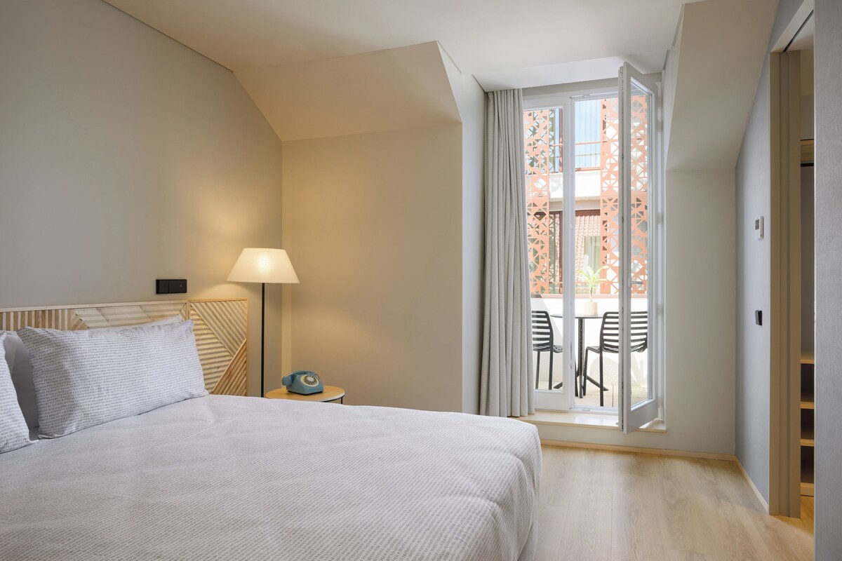 A hotel room with a bed, a nightstand, a lamp, and a view of an outdoor seating area through a glass door.
