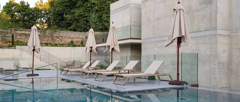 Piscina con sillones de descanso y sombrillas, rodeada por un muro de piedra y árboles.