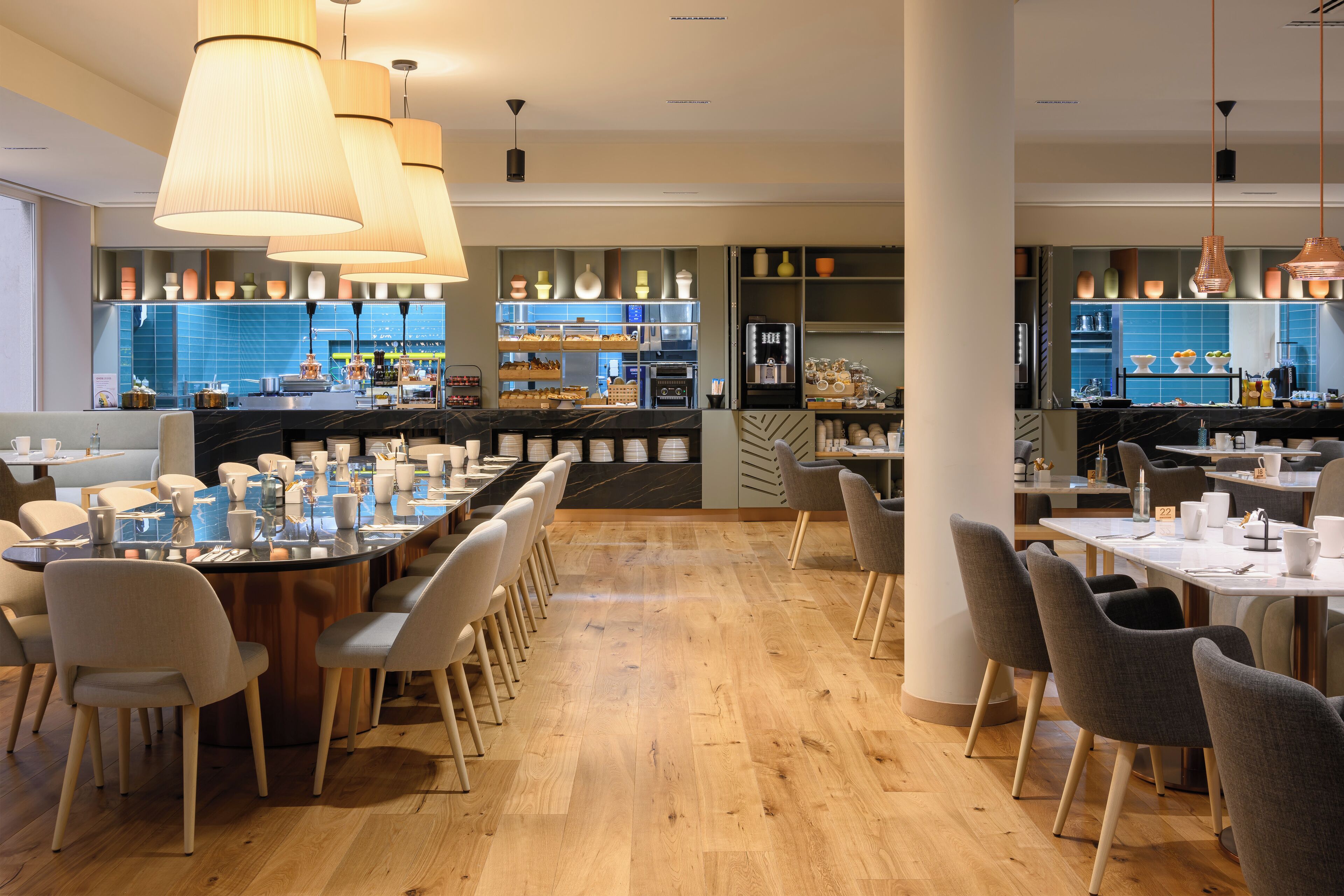 Un restaurant moderne doté d’une grande table conviviale, de tables individuelles et d’un espace cuisine visible au fond de la salle.