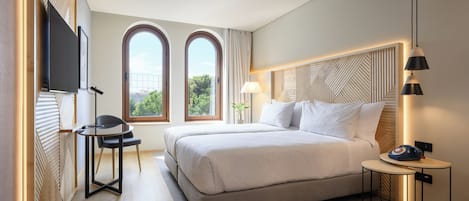 A modern hotel room with a large bed, a desk, a chair, and a TV mounted on the wall.