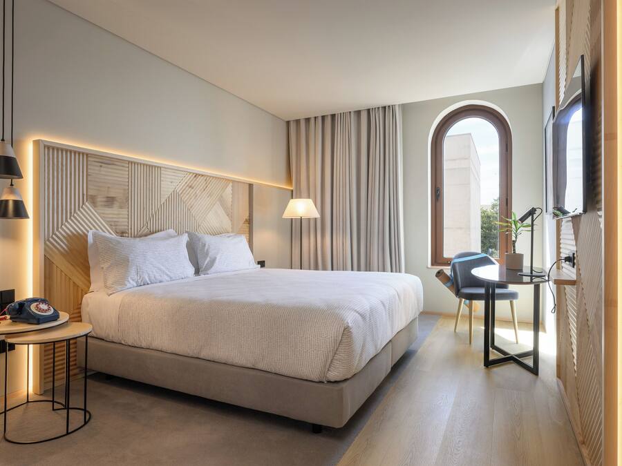 Habitación de hotel moderna con una cama grande, mesita de noche, lámpara y vistas a la ciudad a través de una ventana.