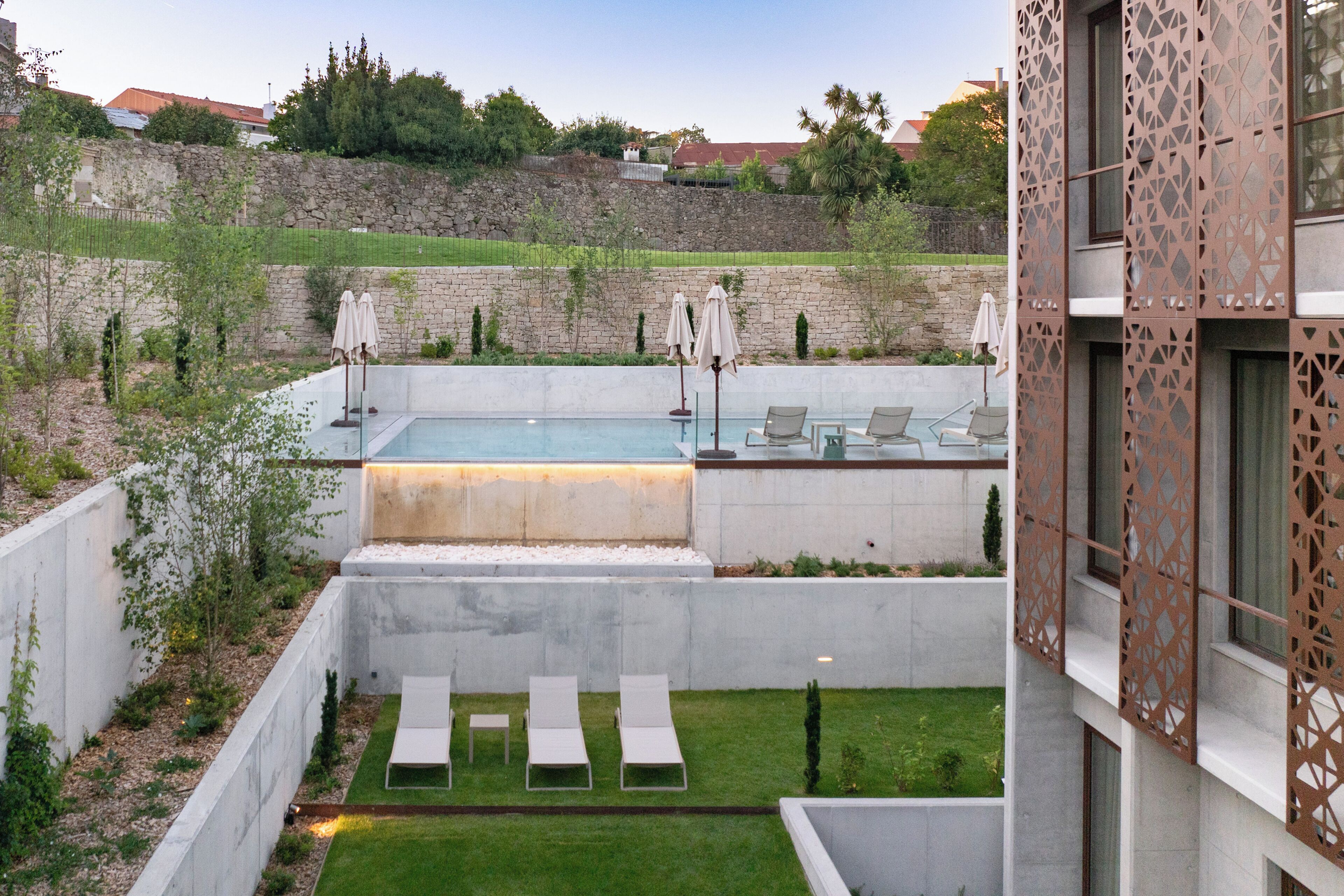 A modern outdoor pool area with lounge chairs and umbrellas, surrounded by a stone wall and greenery.