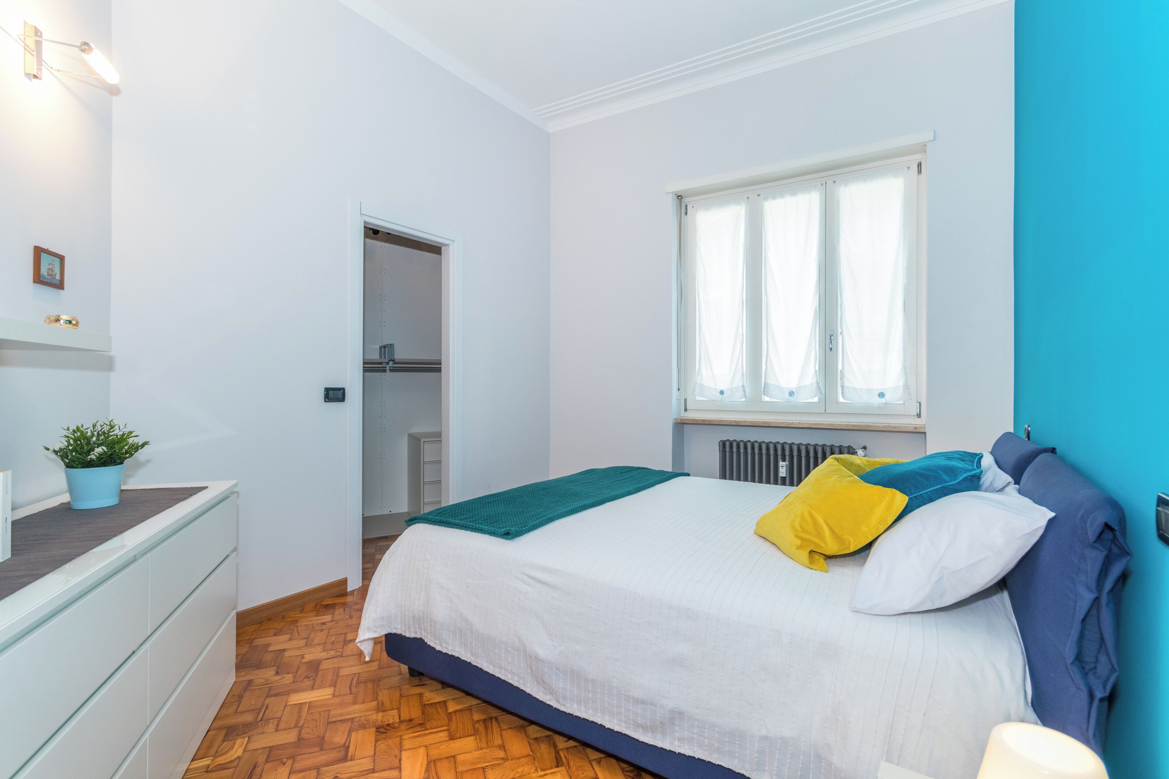 Un dormitorio con una cama, ventana, radiador, puerta y una planta en maceta.