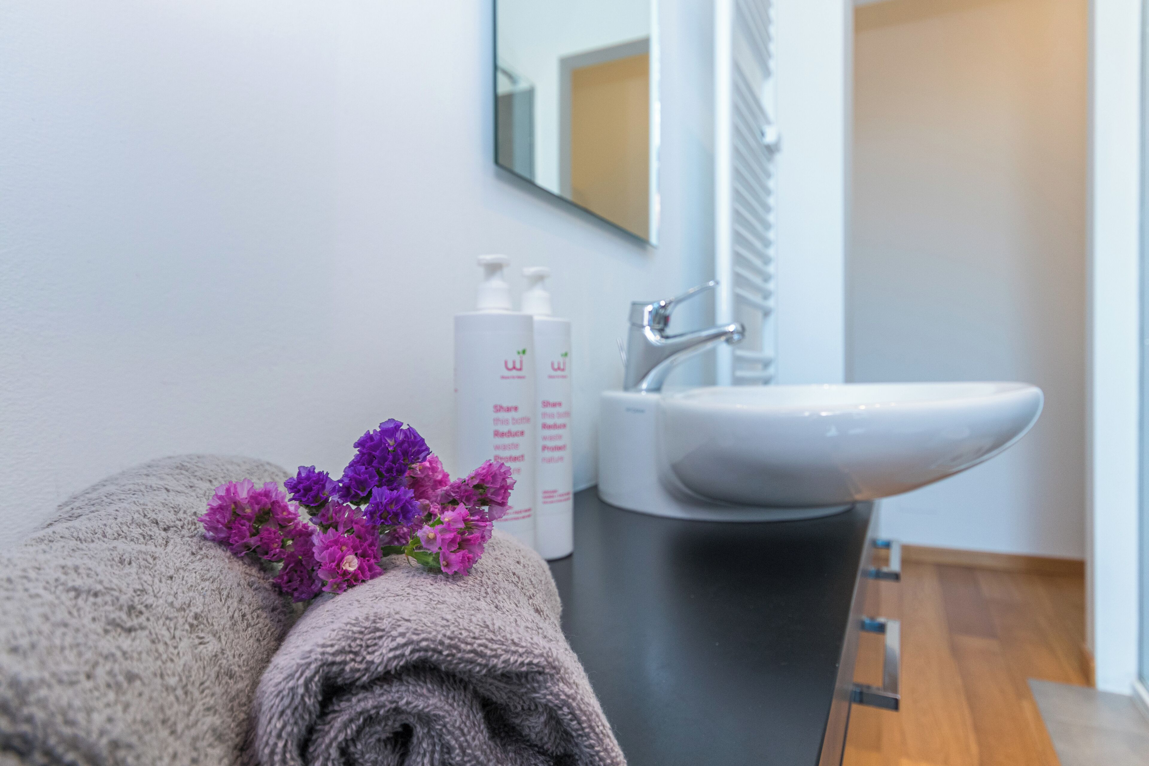 Une salle de bain avec une serviette, des fleurs violettes et des articles de toilette posés sur le comptoir.