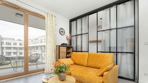 En moderne stue med en sofa, et lille bord med en vase blomster, et ur på væggen og et stort vindue med udsigt til bygninger.