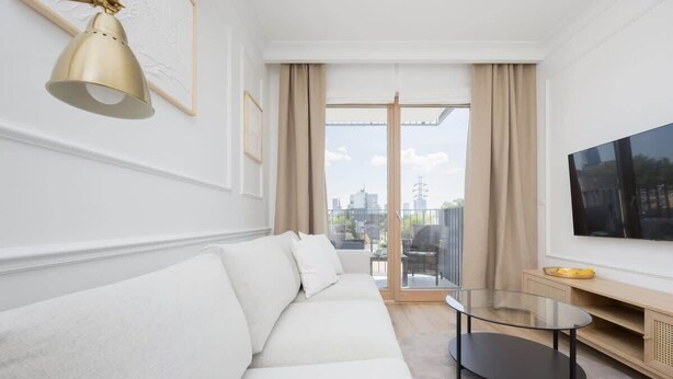 Una sala de estar moderna con un sofá, una mesa de centro de vidrio, un televisor y vistas de la ciudad a través de ventanas amplias.