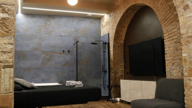 A room with a brick archway, a bed, a TV mounted on the wall, and a wooden floor. - Unique Trastevere (Roma)