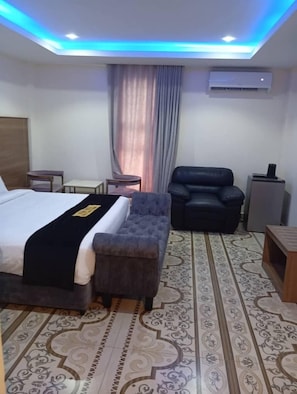 Et hotelværelse med en seng, en grå lænestol, et lille bord, en dekorativ måtte og en aircondition.