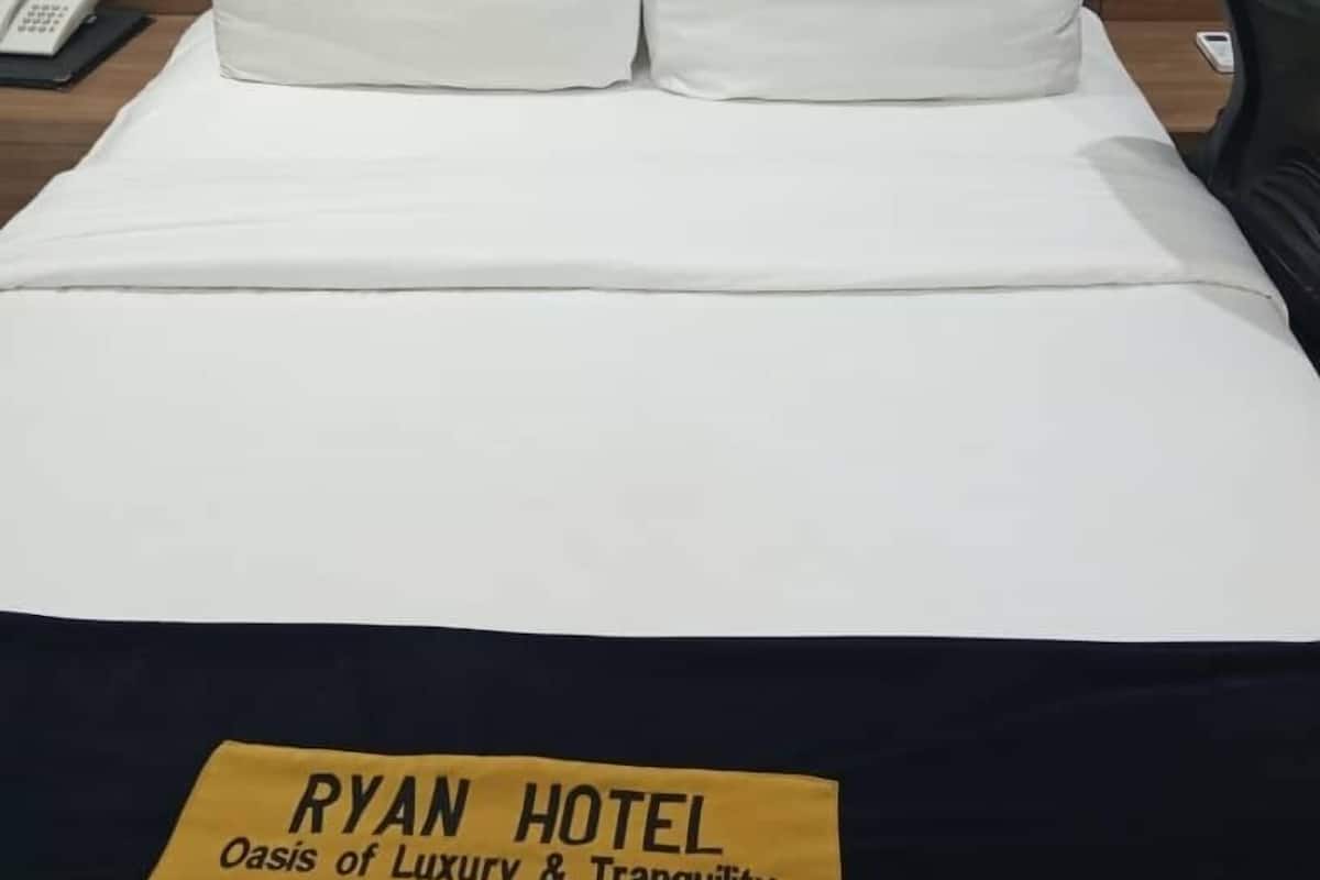 Een netjes opgemaakt bed met witte lakens en een grijs gestoffeerd hoofdeinde. Een geel bord op het bed vermeldt: 'RYAN HOTEL Oasis of Luxury & Tranquility'.