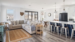 A modern living room with a sectional sofa, a dining area with a table and chairs, a kitchen with a refrigerator and a toaster, and a large window offering a view of the city.