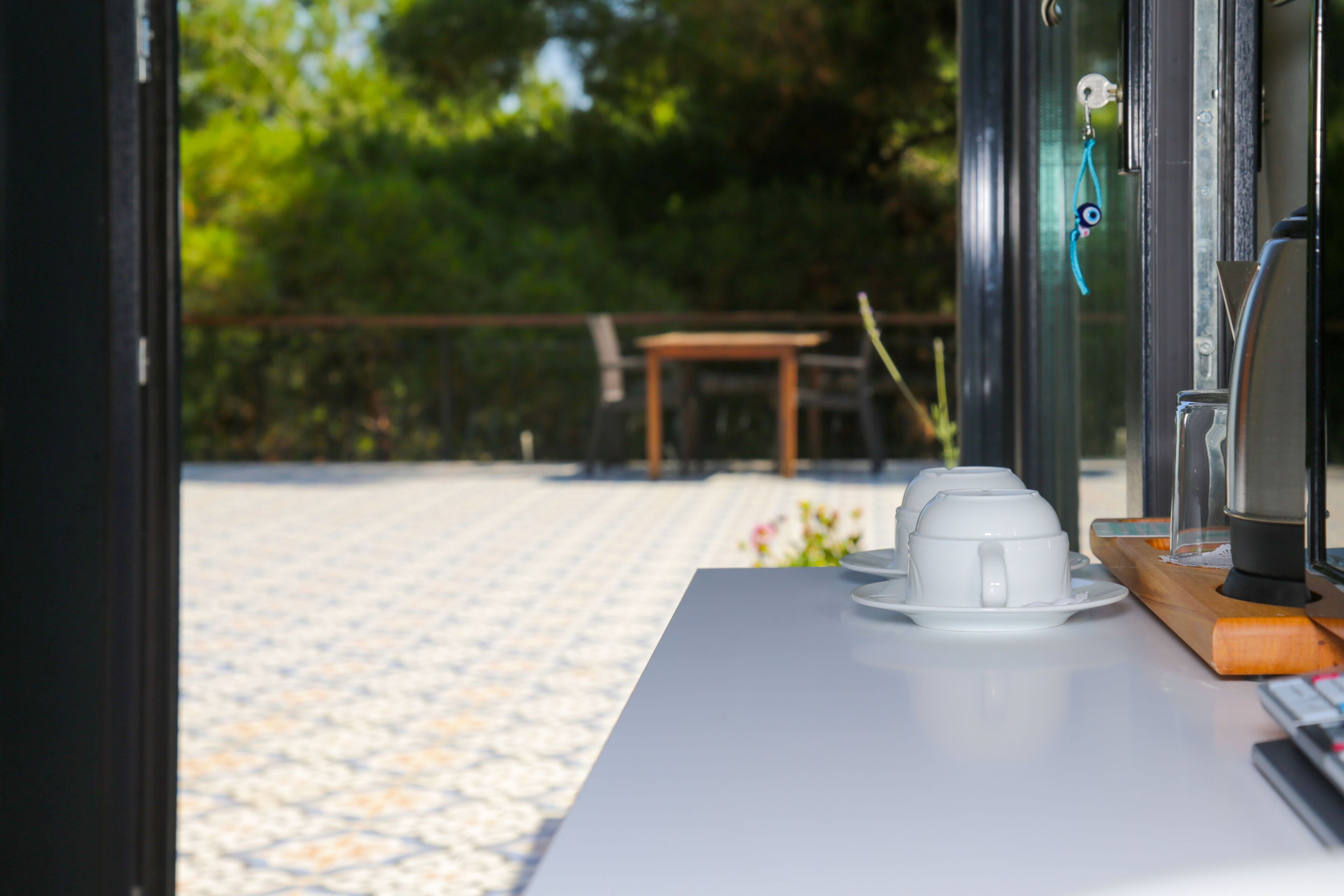 A table with a coffee machine and cups, a wooden tray, and a window with a view of a patio area.