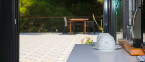 A table with a coffee machine and cups, a wooden tray, and a window with a view of a patio area.