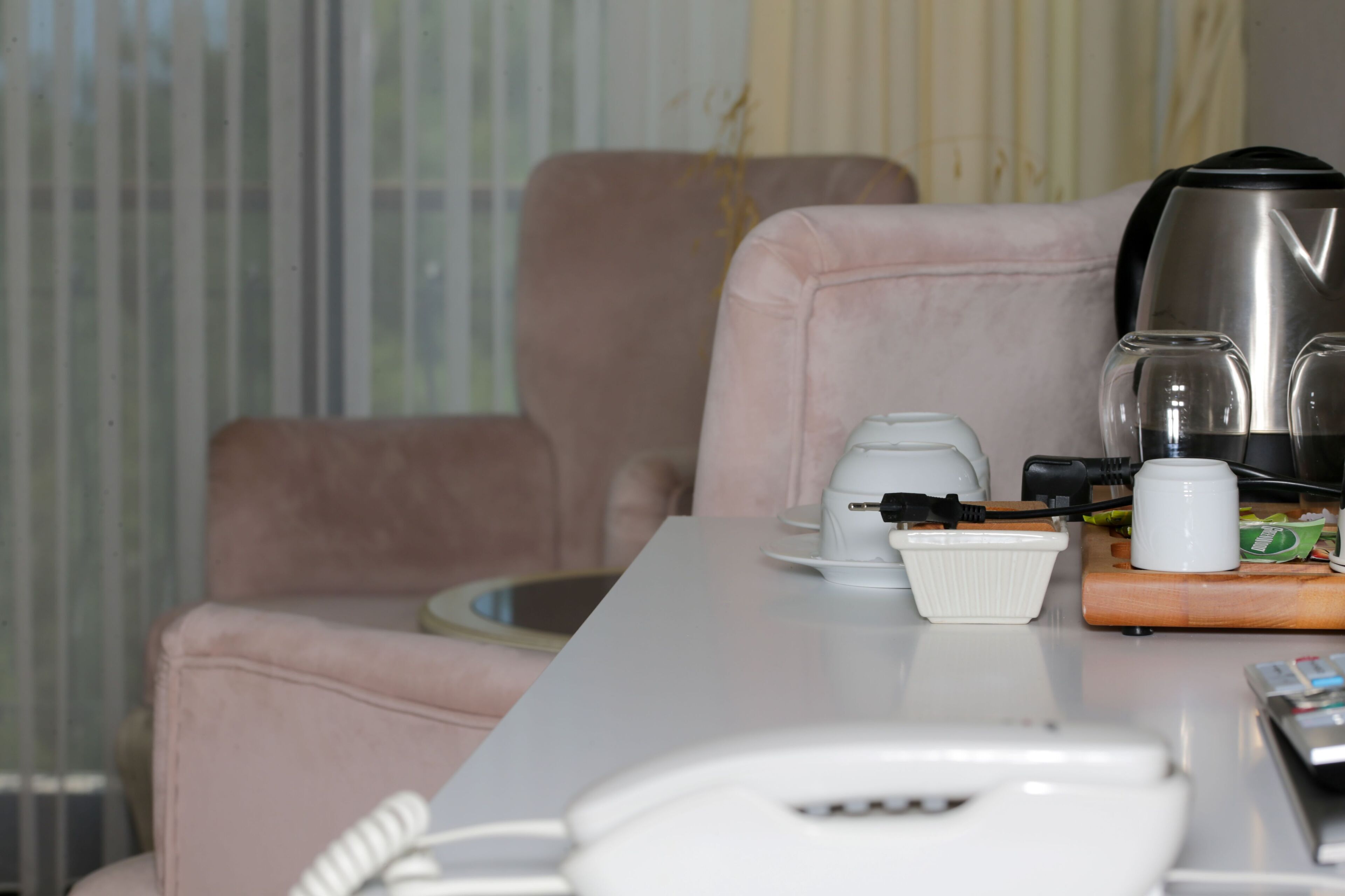 A hotel room with a white table, a pink armchair, a coffee maker, a kettle, and a glass set.