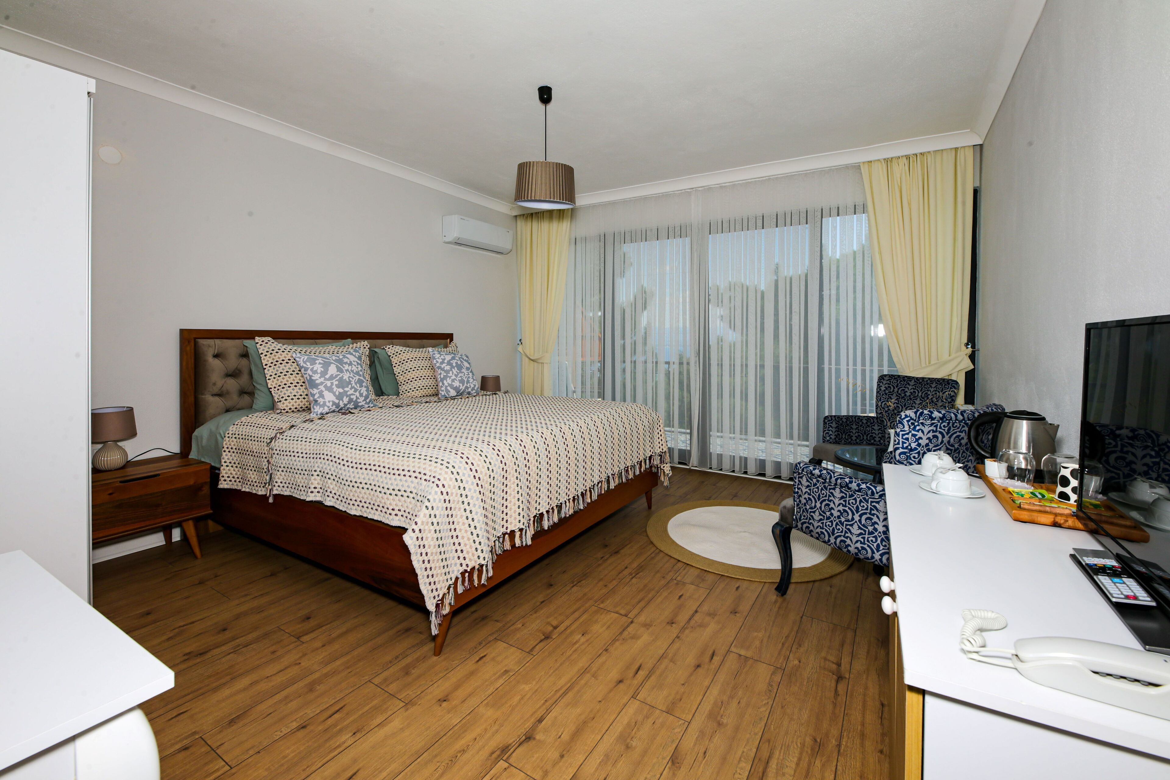 A bedroom with a wooden bed, a bedside table, a desk with a chair, a window with curtains, and a ceiling light.