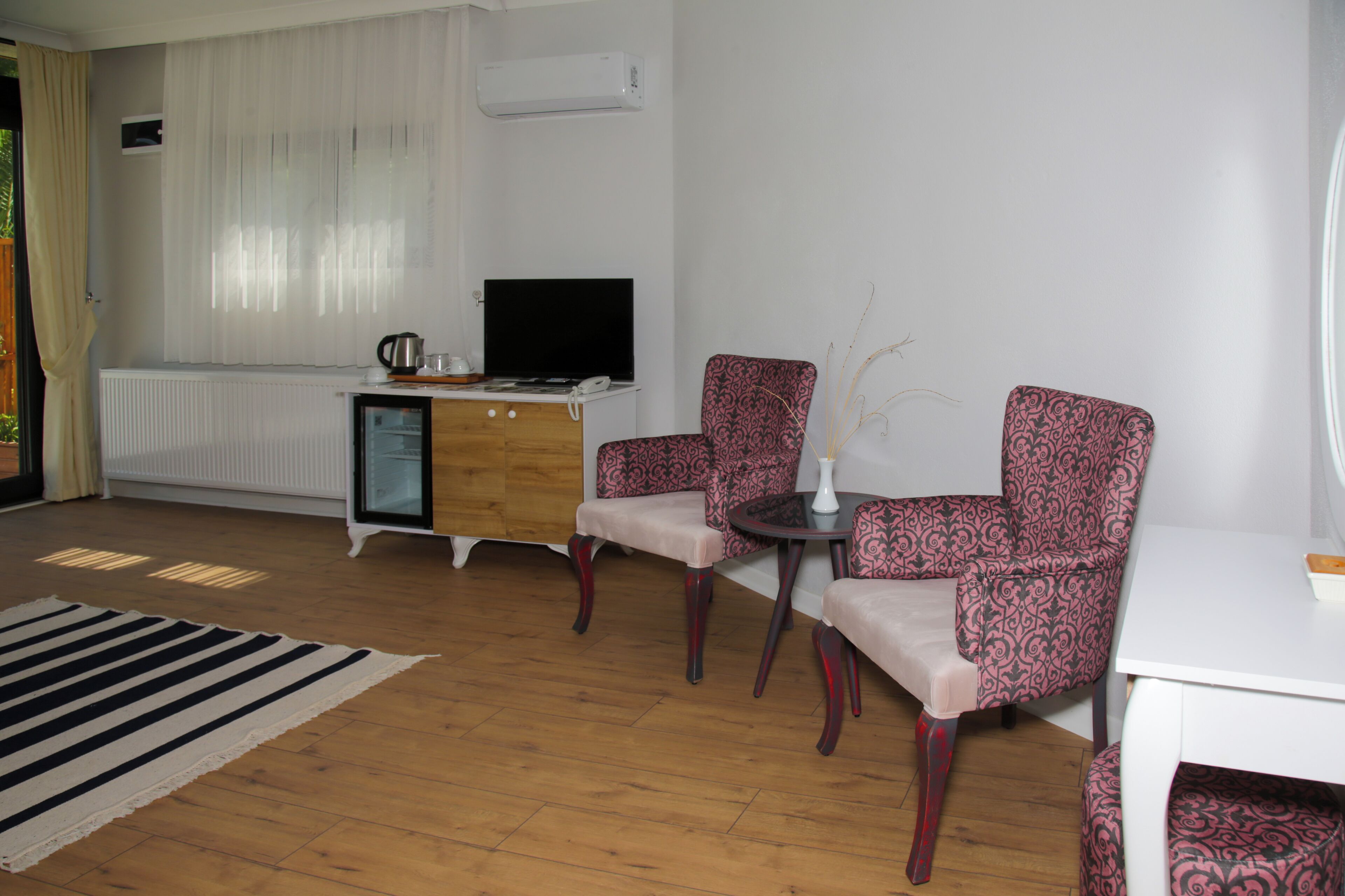 Una sala de estar con piso de madera, un radiador blanco, un televisor, una mesita con un florero y sillas estampadas.