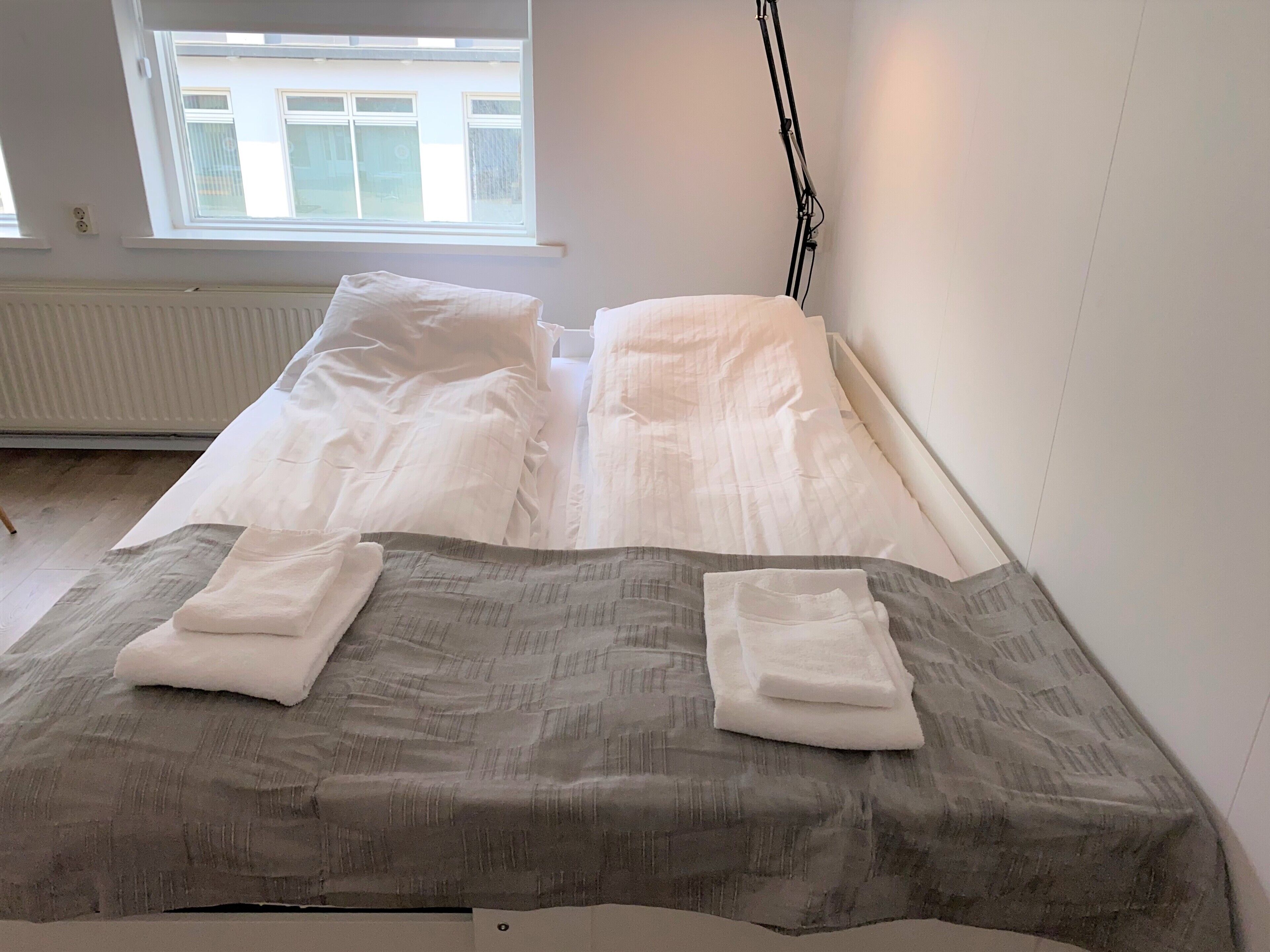 A bed with white linens and a grey patterned blanket, a window with a view of buildings, and a radiator on the wall.