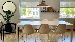 Ein Essbereich mit einem weißen Tisch, vier beigen Stühlen, einer hängenden Pendelleuchte, einem runden Spiegel und einem Blumentopf.