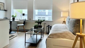 Ein modernes Wohnzimmer mit einer Couch, einem Glastisch, einem Esstisch mit Stühlen und zwei Fenstern mit Jalousien.