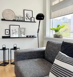 Un salon moderne avec un canapé gris, un coussin à rayures noires et blanches, un lampadaire noir et une fenêtre donnant sur un paysage verdoyant.