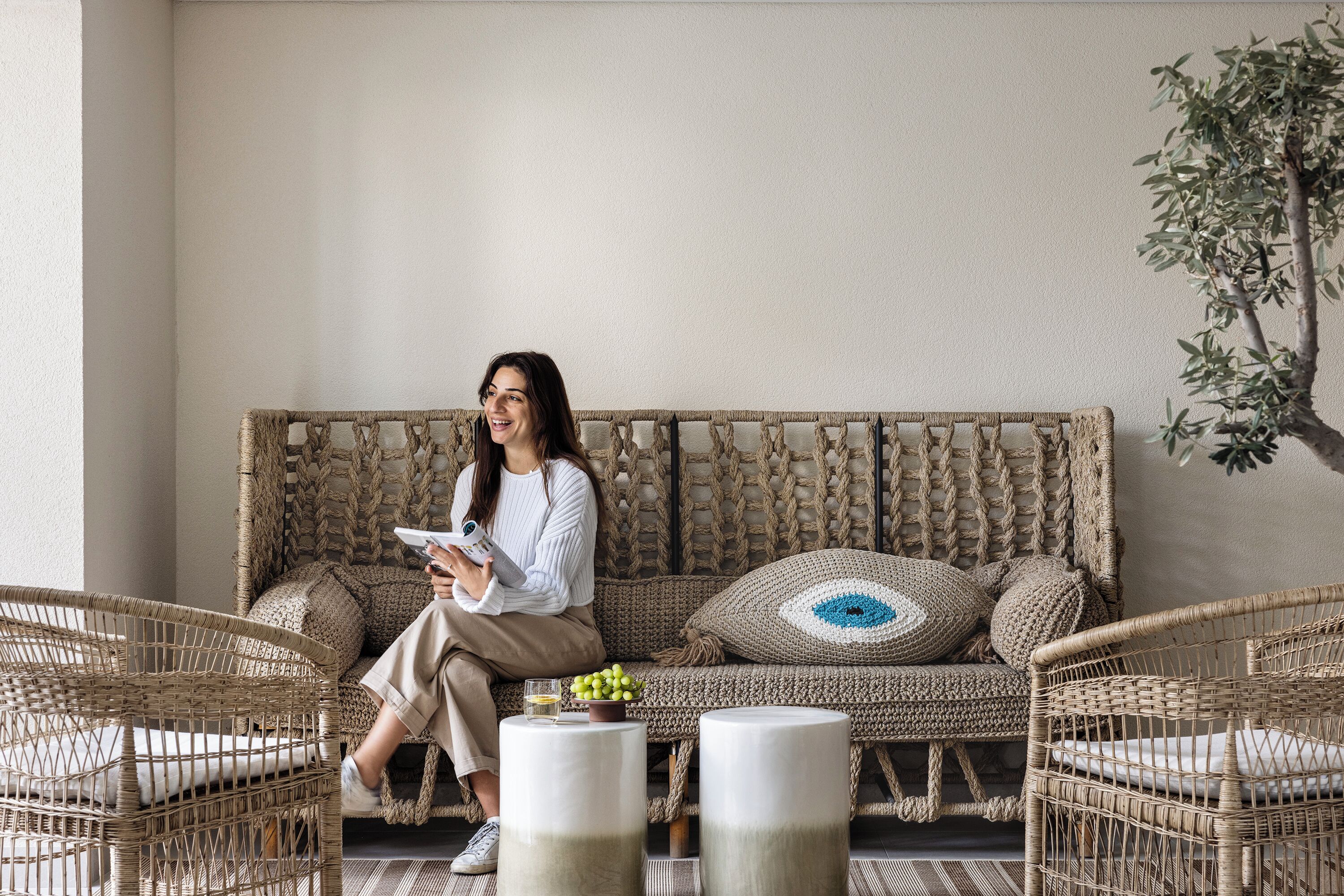 A person sitting on a woven sofa with a view of a tree, surrounded by wicker chairs and a small table with a glass and a bowl of fruit.