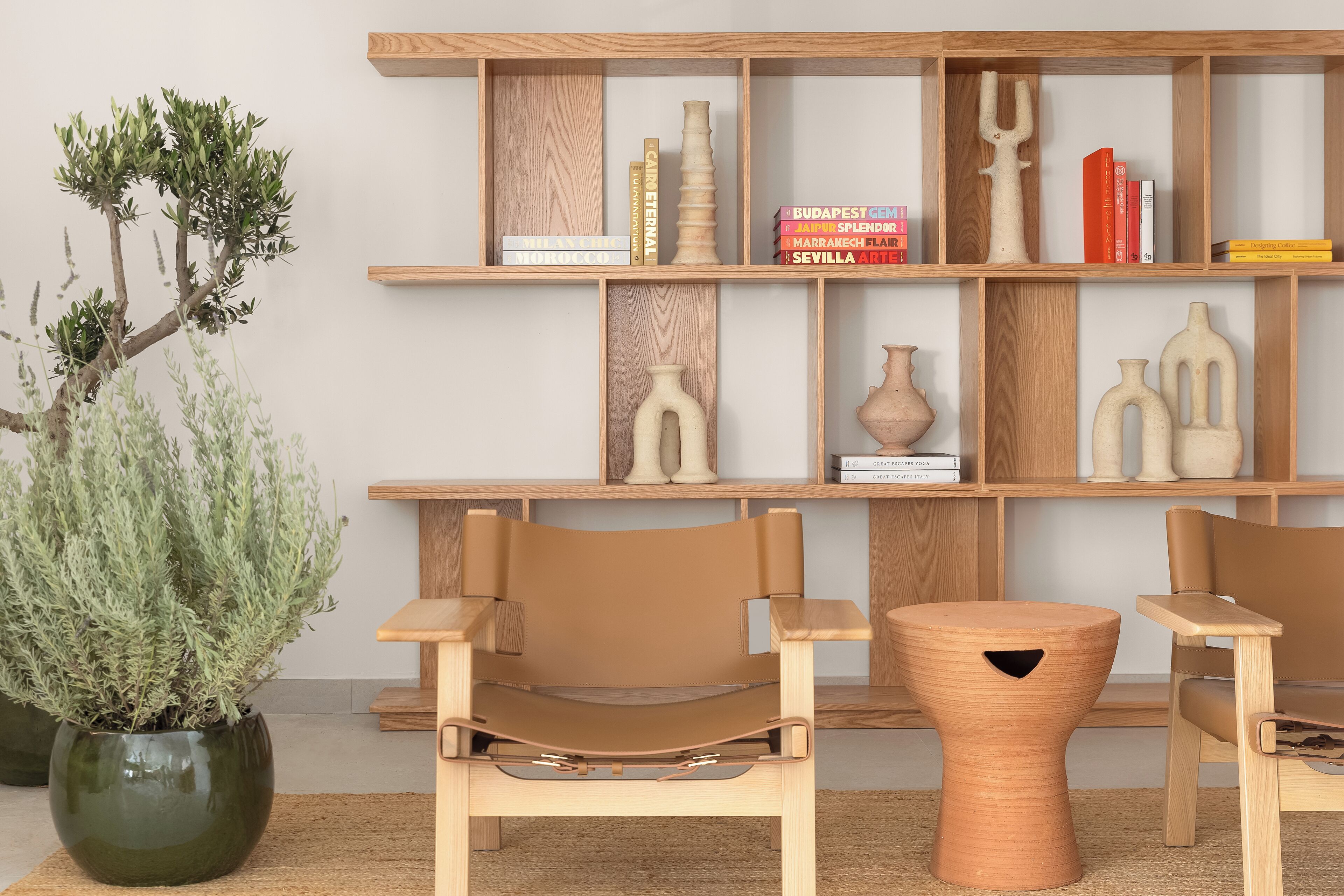 A modern interior with a wooden bookshelf, two leather chairs, a round wooden stool, and a potted plant.