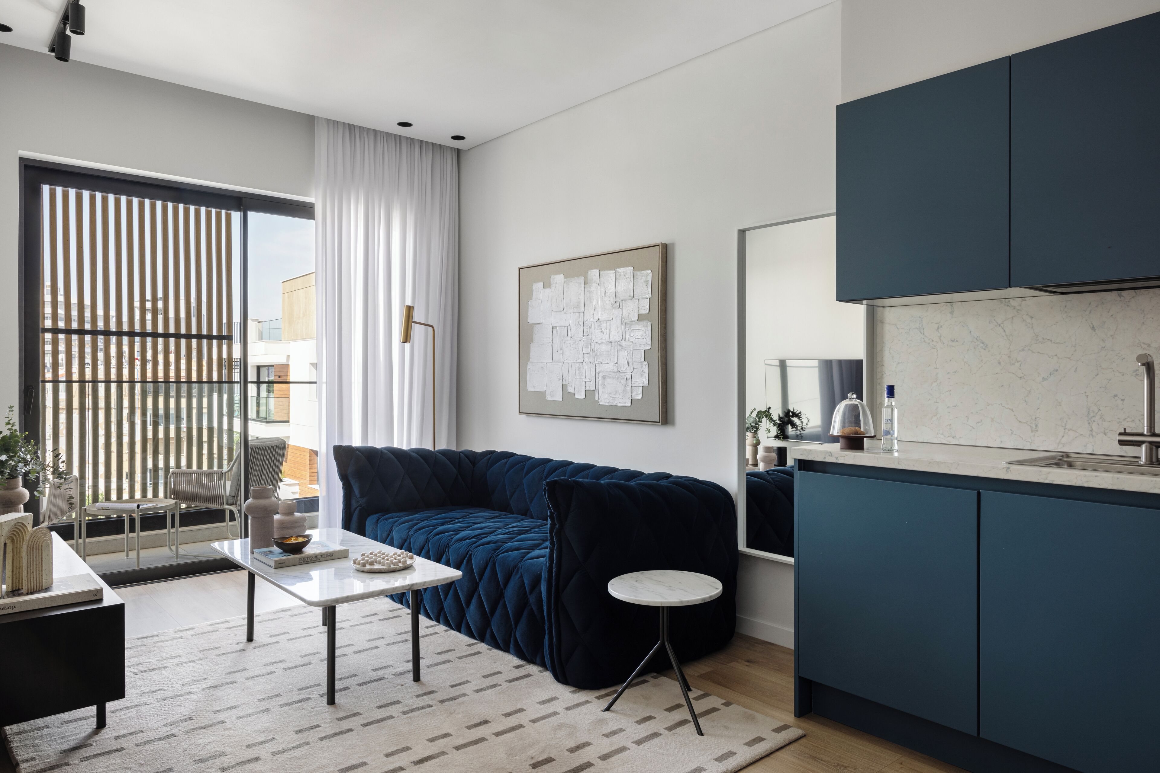 Un salon moderne avec un canapé bleu foncé, une petite table ronde et une table basse. On y trouve une grande fenêtre avec des stores verticaux, un tableau au mur et un coin cuisine avec des meubles bleu foncé et un plan de travail en marbre.