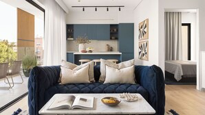 Un soggiorno moderno con un divano blu scuro, un tavolino da caffè con un libro e una ciotola, e vista su una cucina con mobili blu e un'area da pranzo.